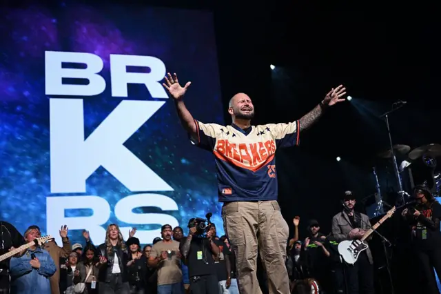 De pé no palco, com os olhos fechados e as mãos estiradas, o pastor Mike Signorelli, vestindo calças bege e camiseta azul, amarela e laranja com a inscrição 'Breakers 25', se dirige a uma conferência no Estado americano de Illinois, em 2025
